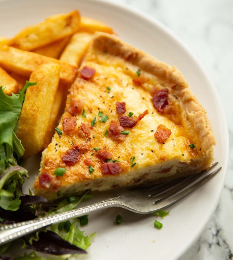 Quiche Lorraine close up shot of slice of quiche lorraine on small white plate with chips and salad
